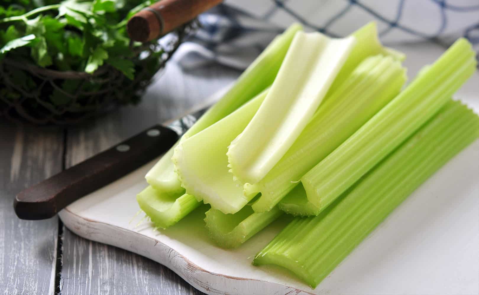 Fresh celery on a white wooden cutting board next to a knife.