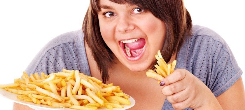 An overweight woman with a plate of fries also holding a handful of fries while looking very eager to eat them.