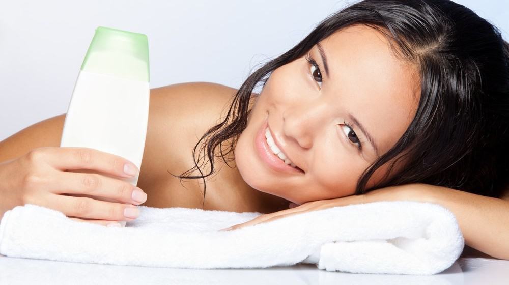 A smiling woman holding a shampoo bottle and lying on a towel.
