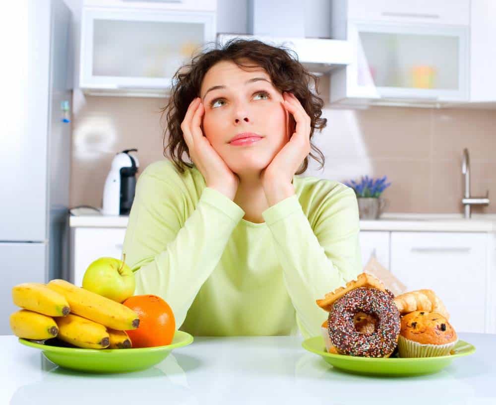 A woman sitting between a bowl of fruit and a bowl of desserts, thinking.