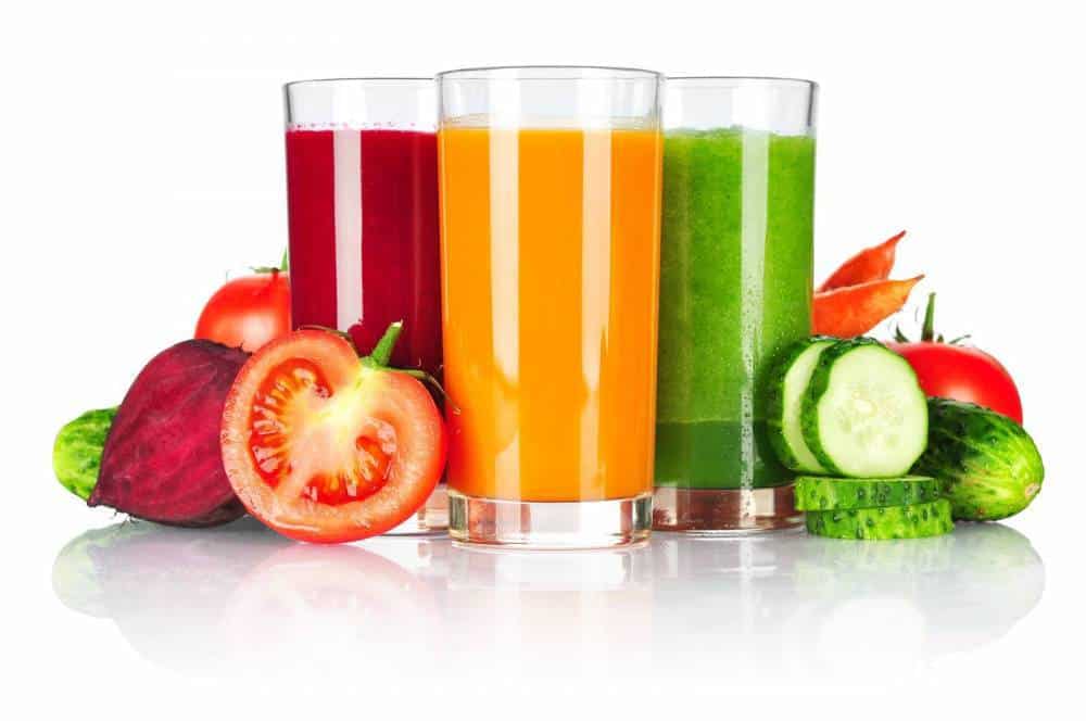 Glasses of vegetable juice surrounded by various vegetables like tomato and cucumber.