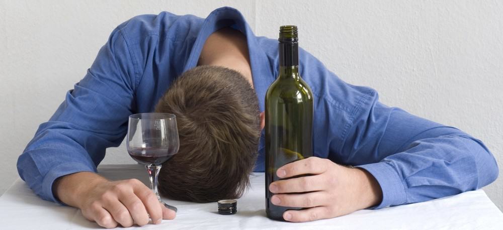 An incredibly drunk man sitting at a table, holding a bottle of wine and a glass with his head held down.