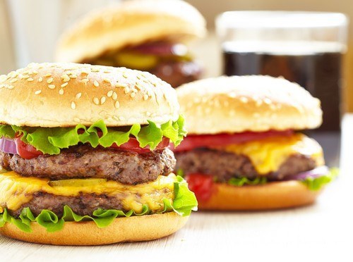 A juicy hamburger with two others burgers and a glass of soda in the background.