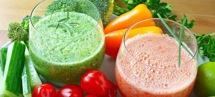 Red and green vegetable smoothies surrounded by carrots, cucumbers and tomatoes.