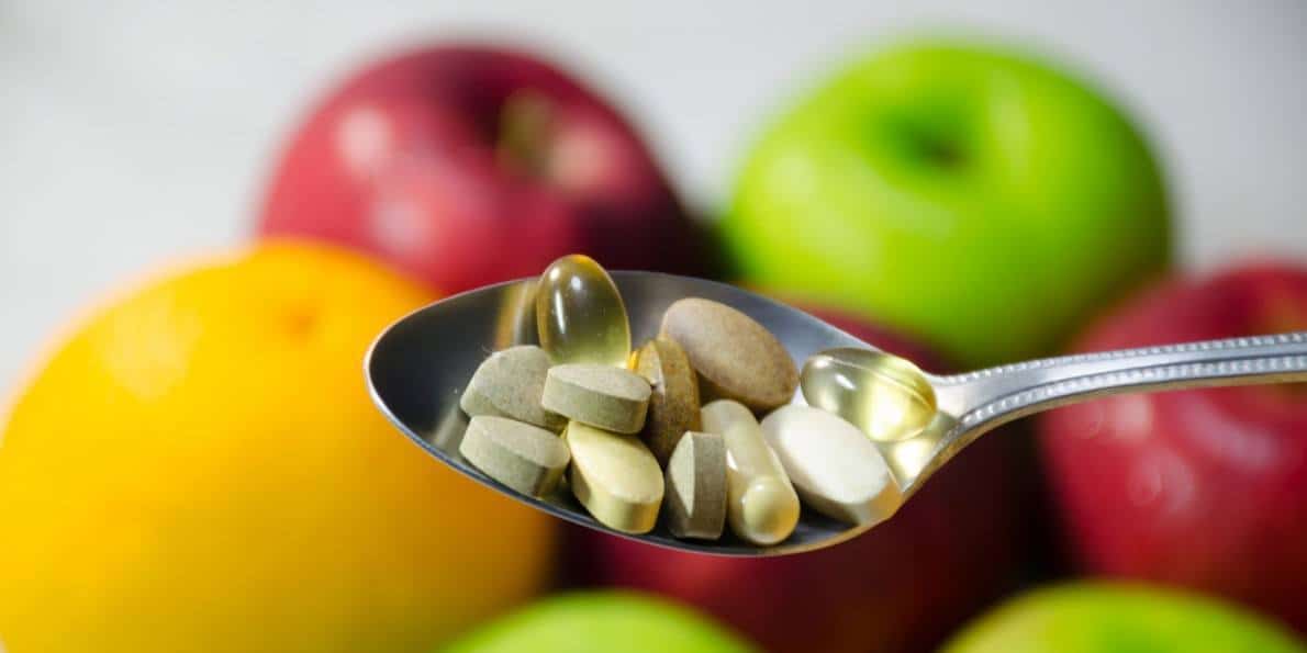 A spoonful of vitamin capsules in front of a background of fruit.