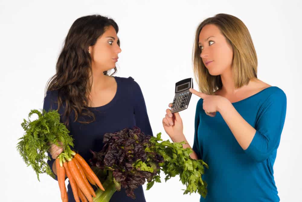 A woman showing a calculator to another woman holding vegetables.