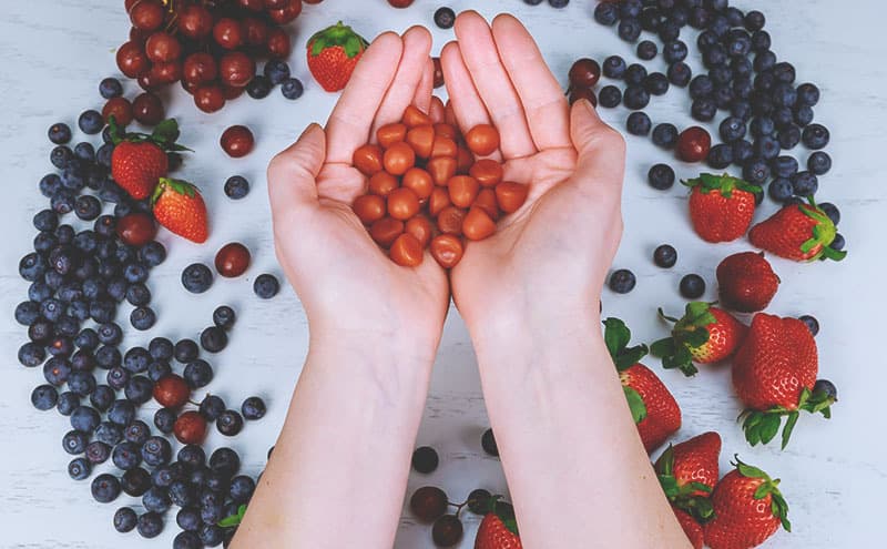 Two hands holding pieces of the best gummy multivitamin above different kinds of berries on a light background.