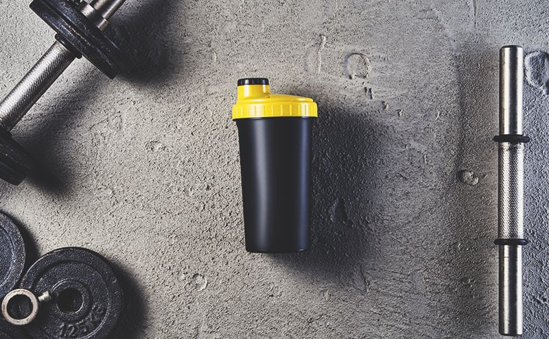 One of the best protein shakers bottles surrounded by a dumbbell, wights and a dumbbell bar, on limestone background.