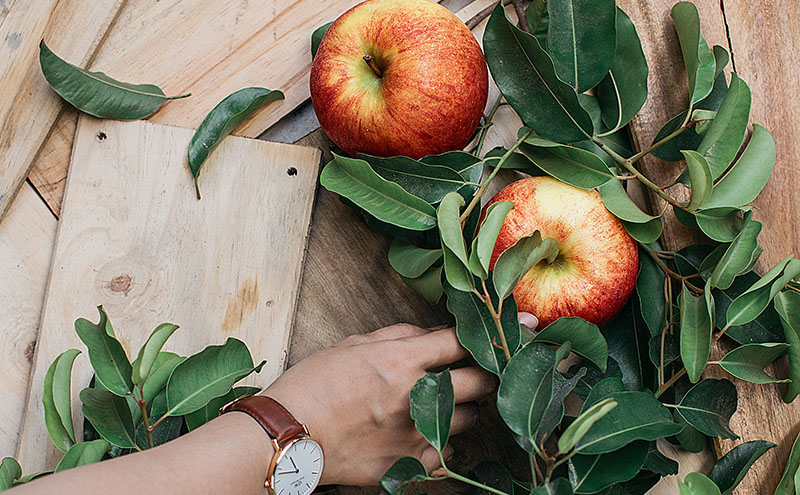 Two apples and multiple leaves from an apple thing on a light wooden boards and a hand with a watch on its wrist reaching for one of the apples.