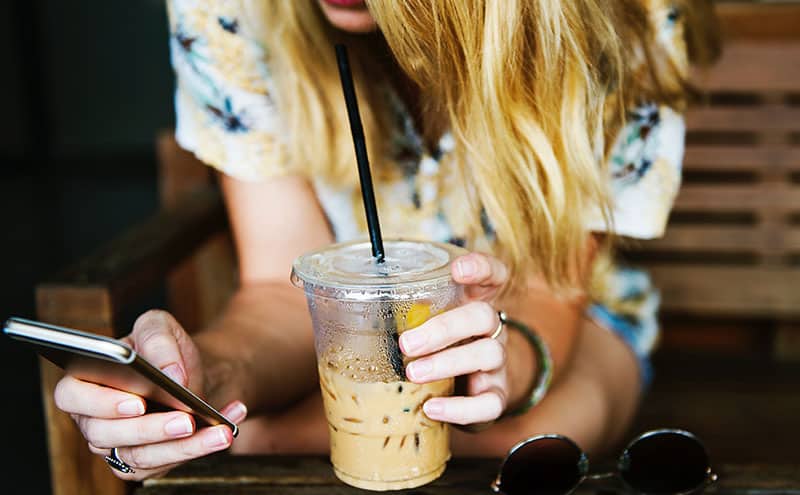 Woman with ice coffee in her hand, looking at her phone, trying to find out if caffeine addiction is a myth or reality on the internet.