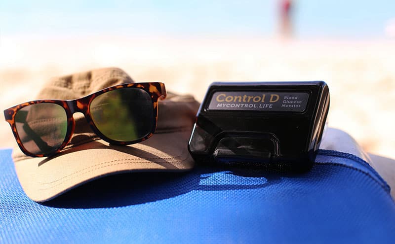 A cap, sunglasses and a blood glucose monitor at what appears to be a beach, while woman is getting ready to check blood sugar, after consuming fructose.