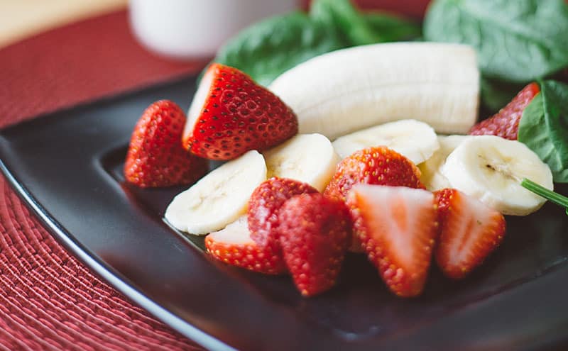Strawberries, a half and sliced bananas, leaves on a dark square-shaped plate.