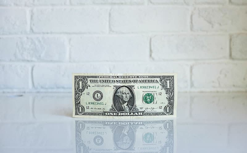 A one dollar bill standing on its side on a reflective white surface, in front of a white brick wall.