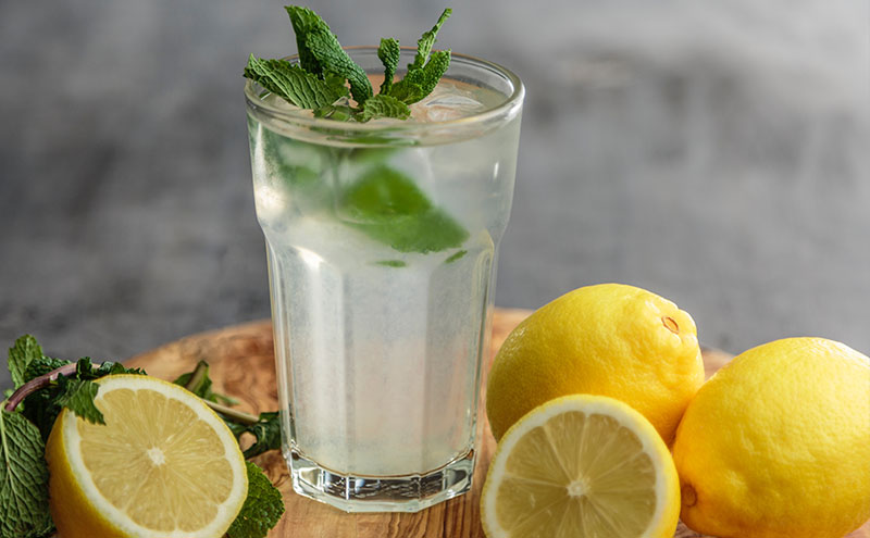 A glass of lemon water with ice cubes and leafs of mint, surrounded wit two full two half lemons on a small medium brown wooden table.