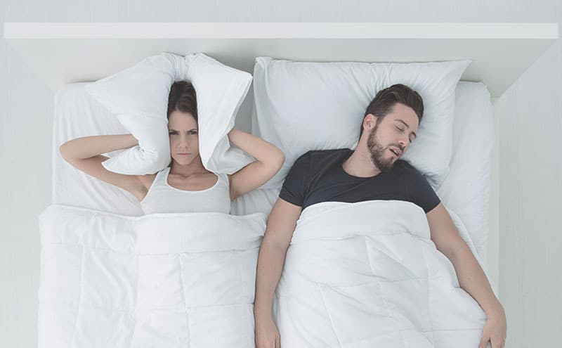 Woman covers her ears with her pillow because of her snoring husband in the bed.