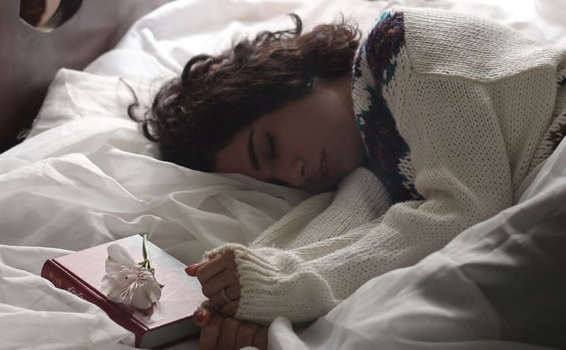 Woman in sweater sleeping on her side in her bed, with a book and a flower next to her.