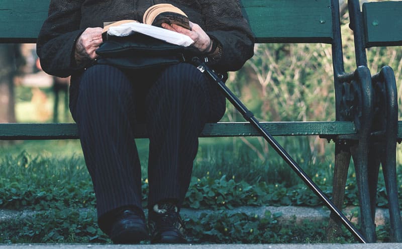An old lady reading a book on a bench while holding her collapsible walking cane